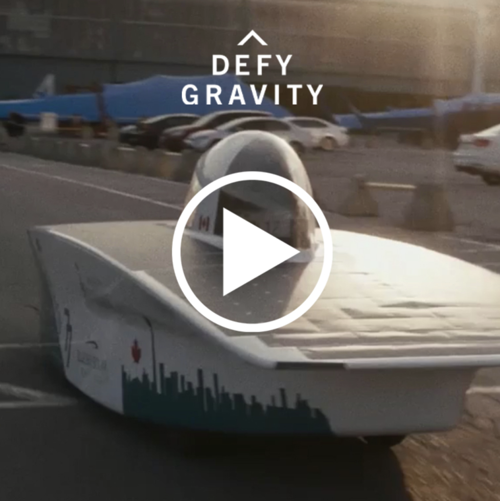 The U of T Blue Sky Solar Racing car, driving in a parking lot, is low to the ground with a flat top covered in solar panels 