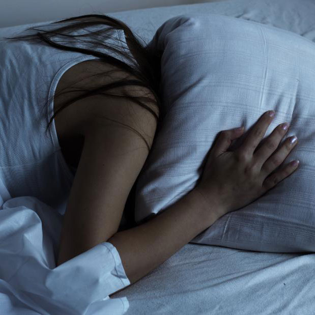 A woman lies face down in bed and holds a pillow over top her head at night