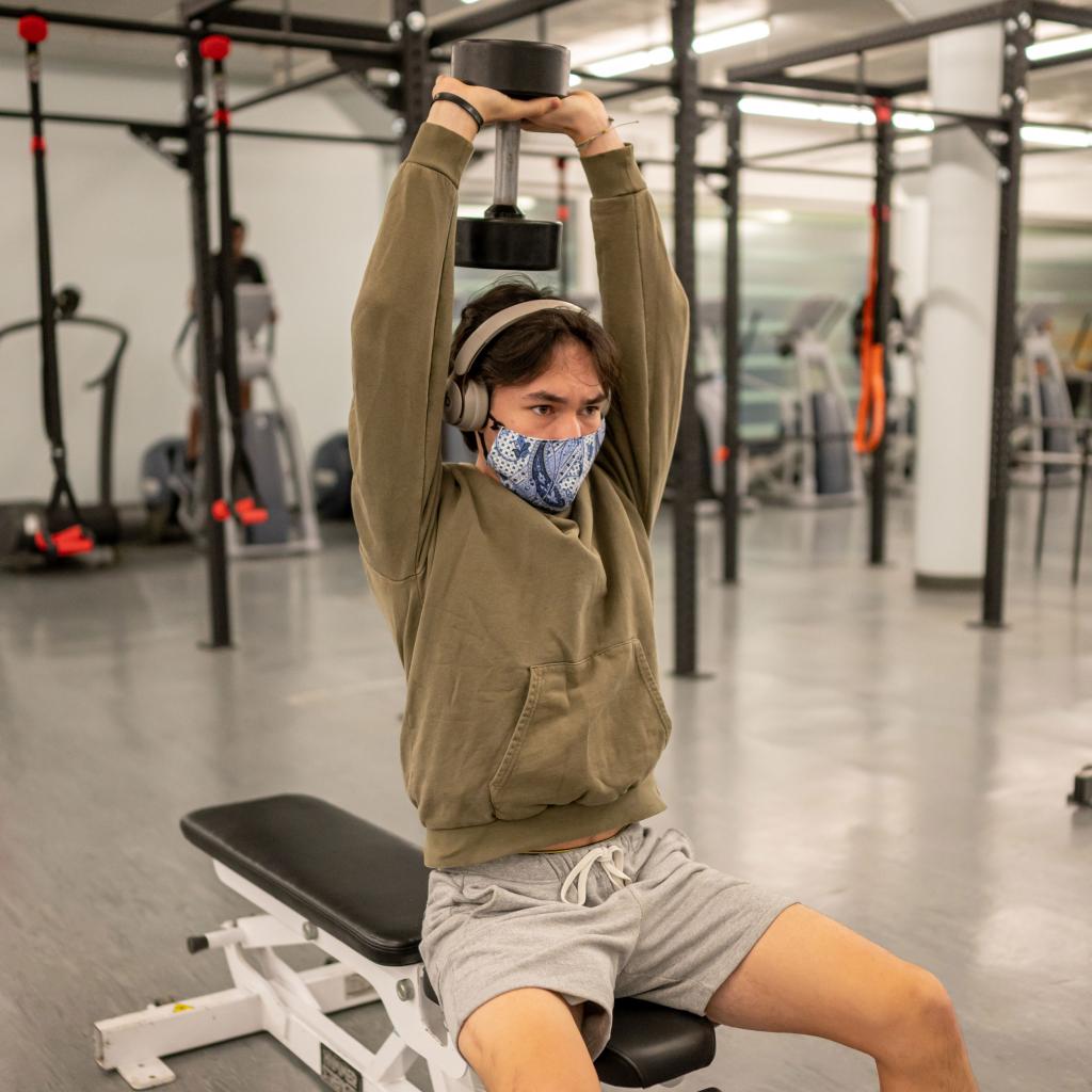 An Asian man wearing a face mask sits on a workout bench. He holds a dumbbell over his head. 