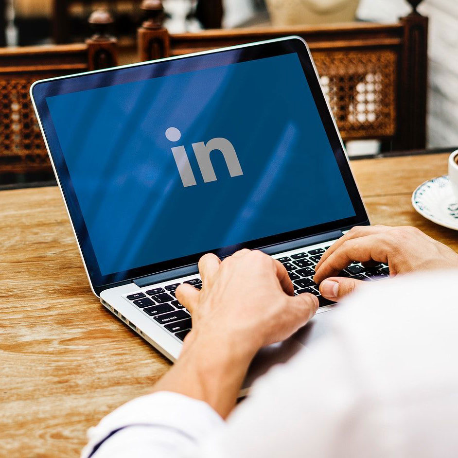 A man sits at a table using a laptop. The LinkedIn logo is featured on the computer screen