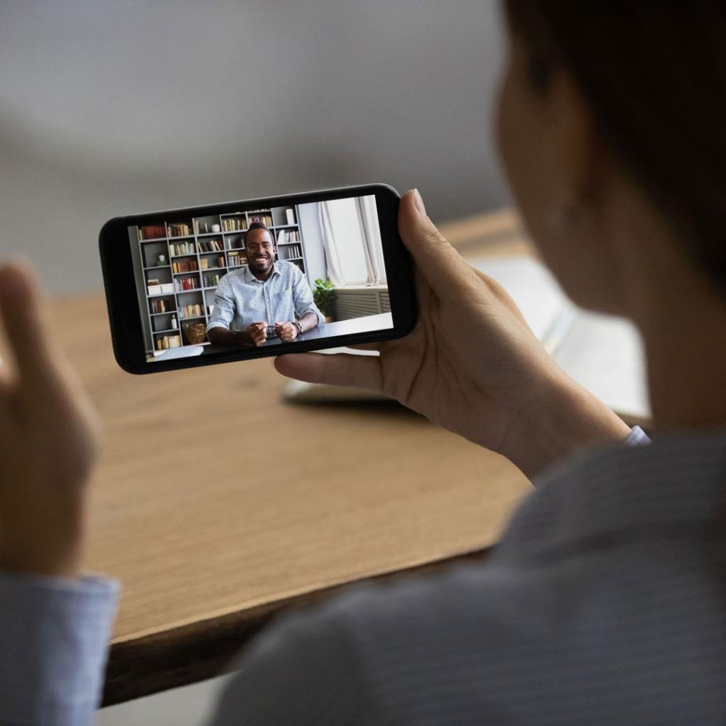 A woman sits at a desk taking a video call via a mobile phone. A man also sitting at a desk is shown on screen smiling 