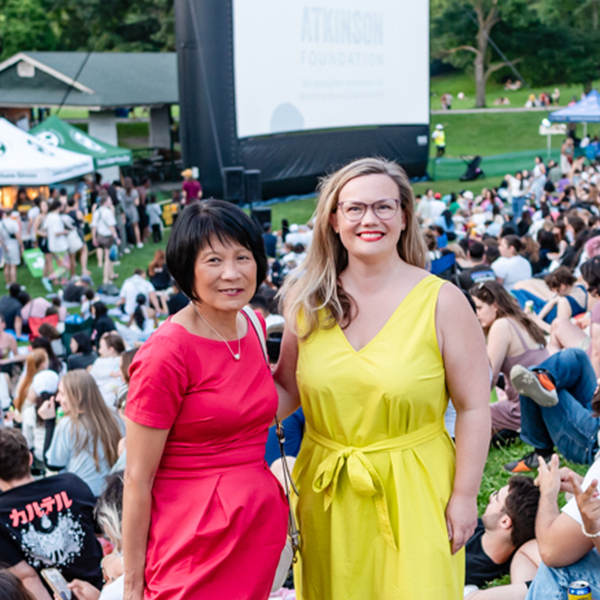 A woman in a red dress stands with a woman in a yellow dress amid a crowd of seated people.