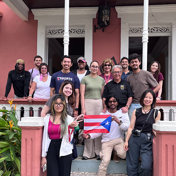 A group of smiling people stand on a porch with two people holding a Puerto Rican flag.