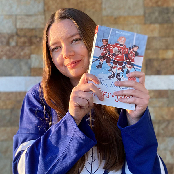 A woman with brown hair and wearing a Toronto Maple Leafs jersey holds up a children's book.