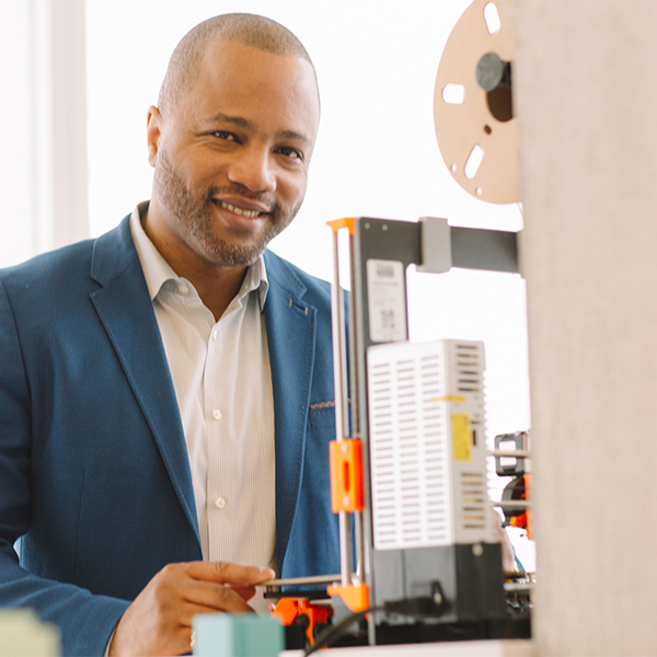 A man with a trim beard and blue suit jacket smiles near technology.
